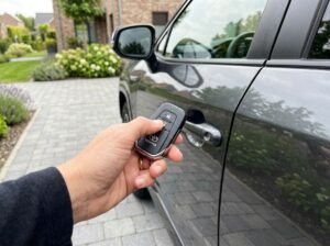 Hand holding key fob remote keyless entry near parked car in driveway